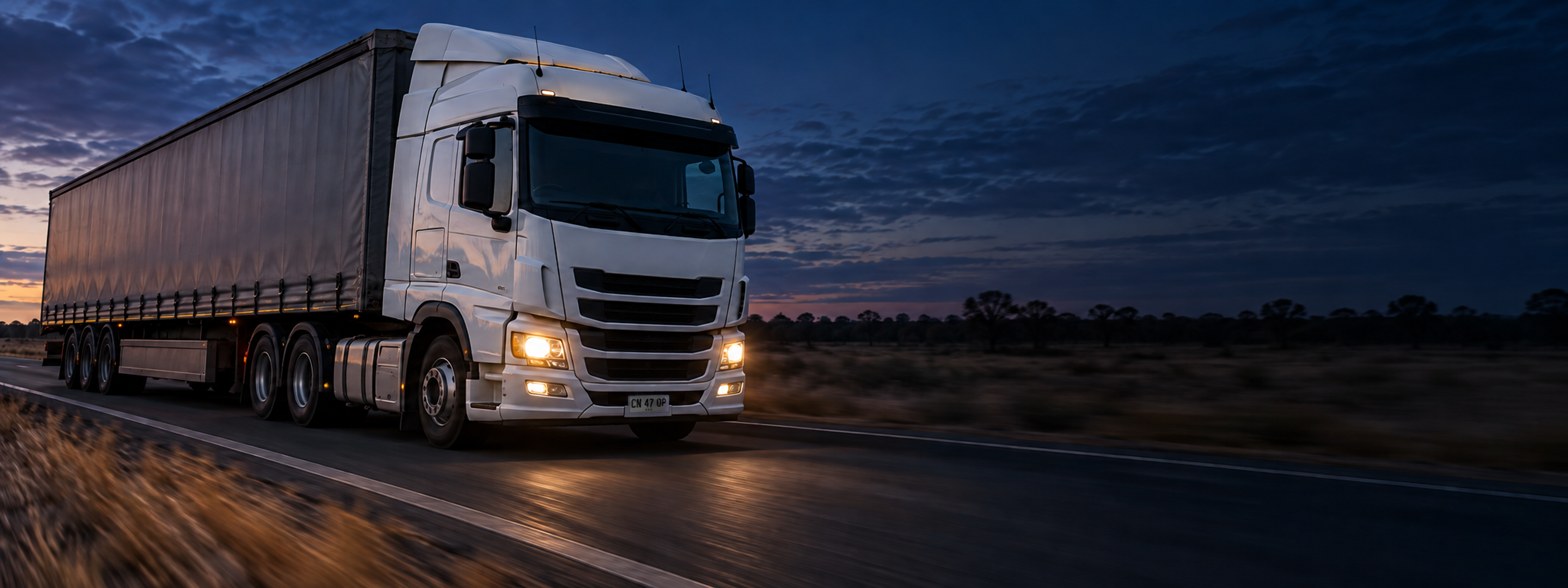 A curtain-sider on a highway at speed, shot from the front three-quarter with motion blur behind. Dark and cool enough for a panel to sit over the right third. No text in the artwork. 2400x900.