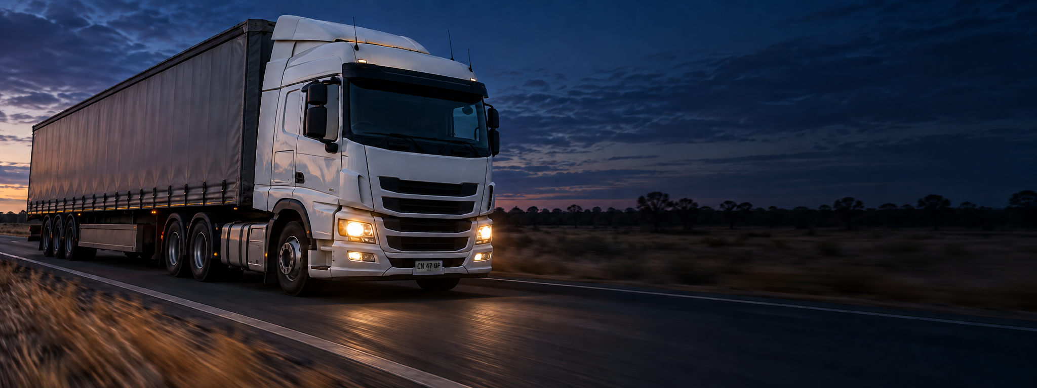 A curtain-sider on a highway at speed, shot from the front three-quarter with motion blur behind. Dark and cool enough for a panel to sit over the right third. No text in the artwork. 2400x900.