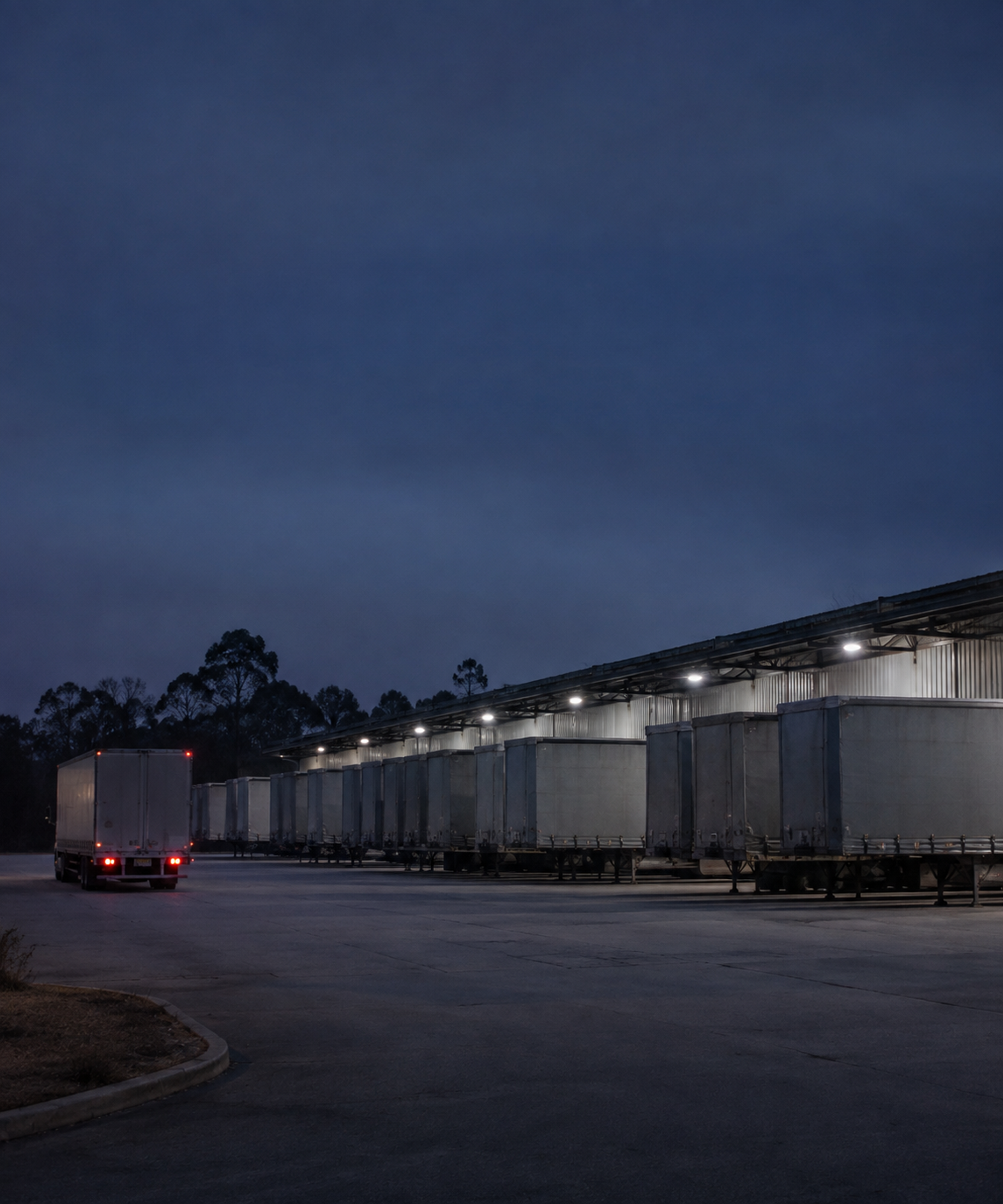 Photograph: a distribution yard before dawn, trailers under lights, one prime mover pulling out. Dark and cool, room at the top, no text in the artwork.