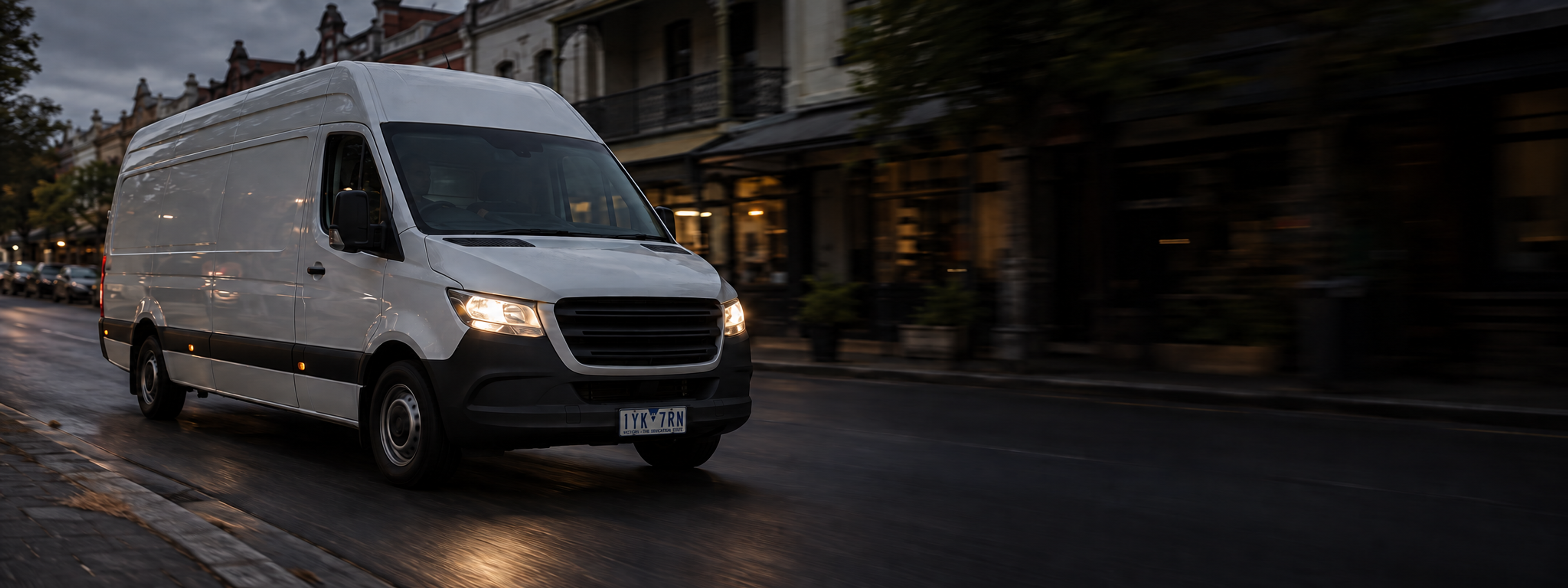 A delivery van on a city street in daylight, shot from the front three-quarter. No visible manufacturer badge. Dark and cool enough for a panel to sit over the right third. No text in the artwork. 2400x900.
