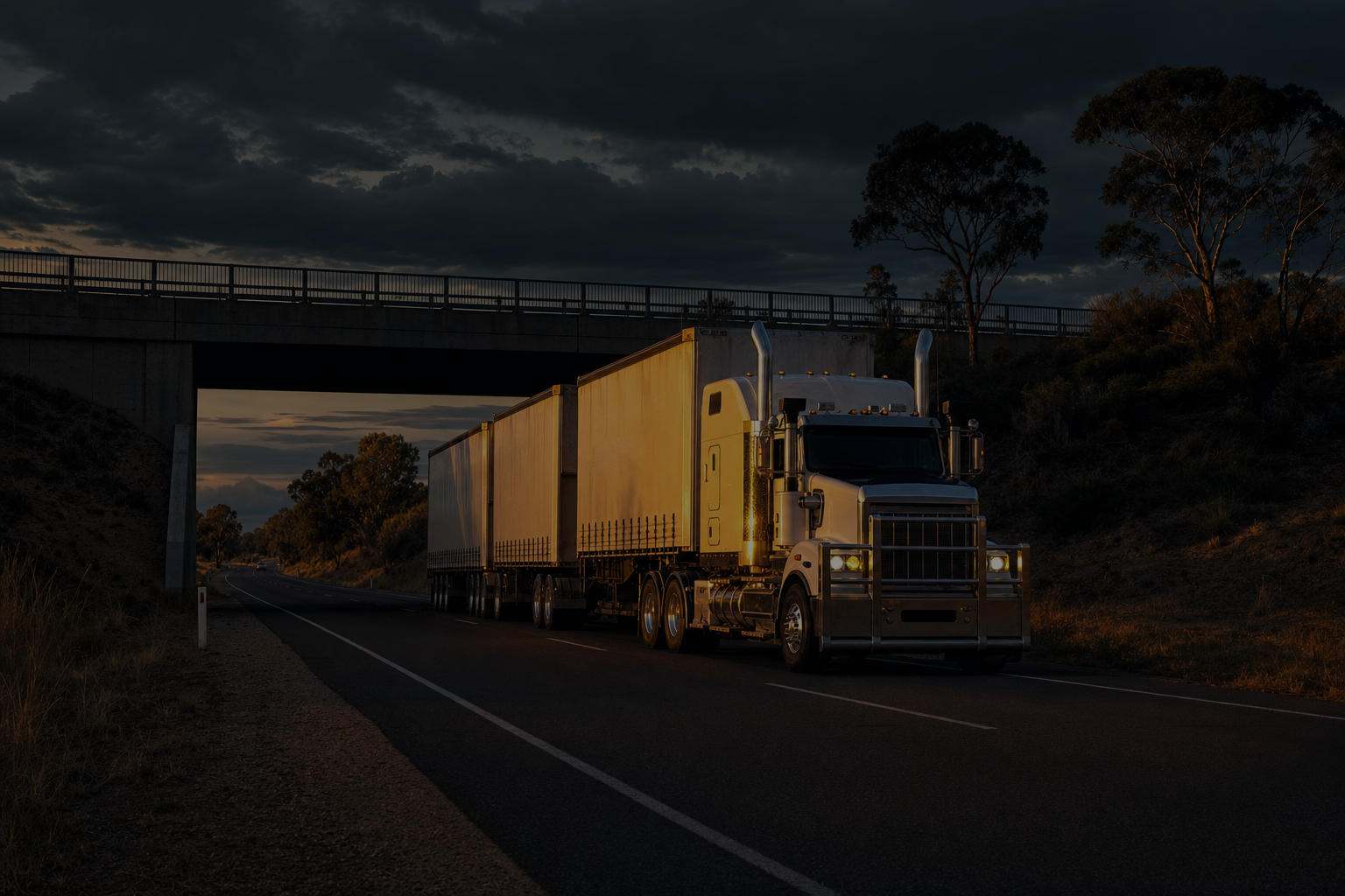 Full-bleed scene, edge to edge: a B-double on a rural highway at golden hour approaching a low bridge, dark and cool so the headline and the accordion read over it. No text in the artwork. 2400x1400.