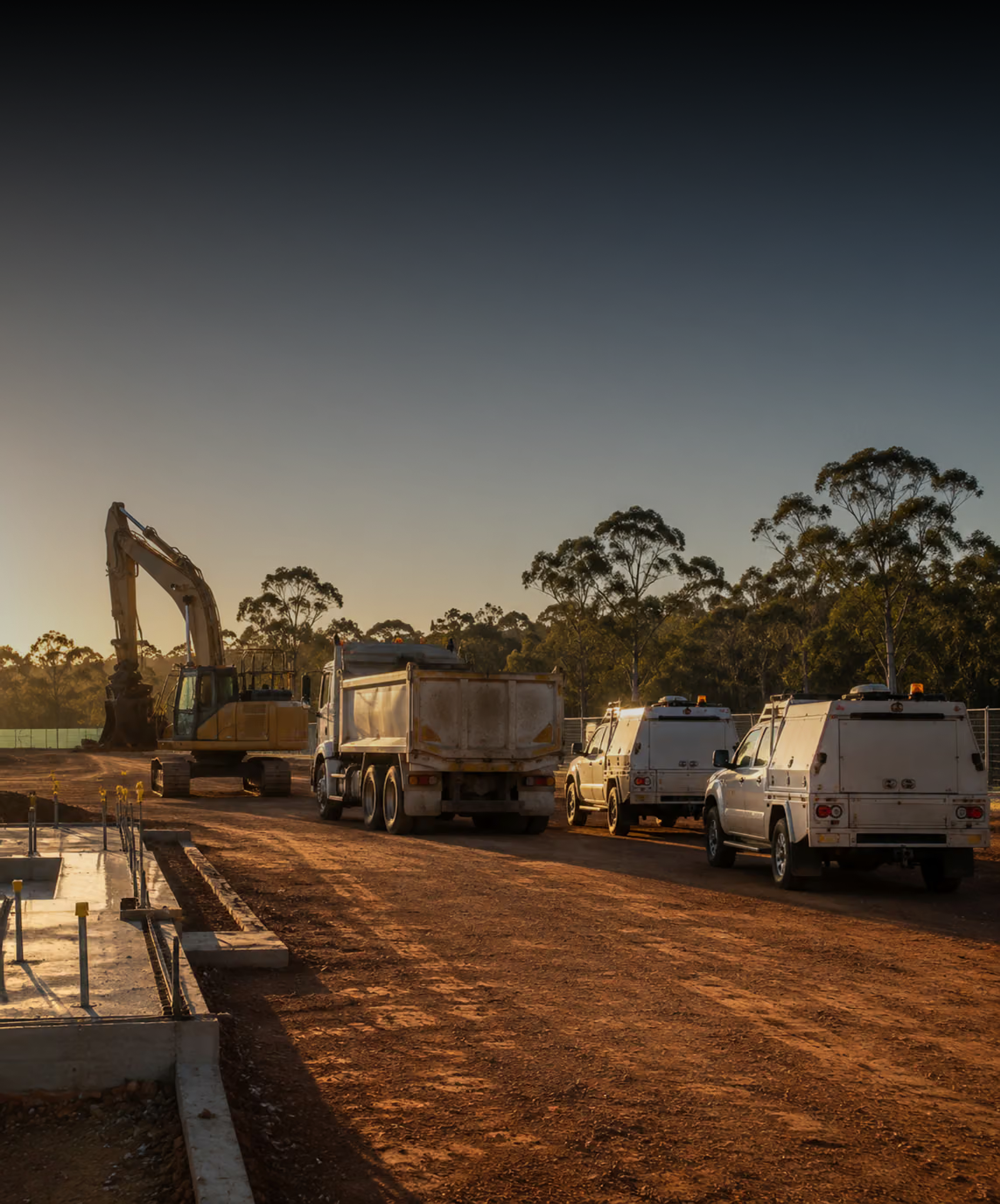 Photograph: a site fleet on a working construction site at first light. Dark and cool, room at the top, no text in the artwork.