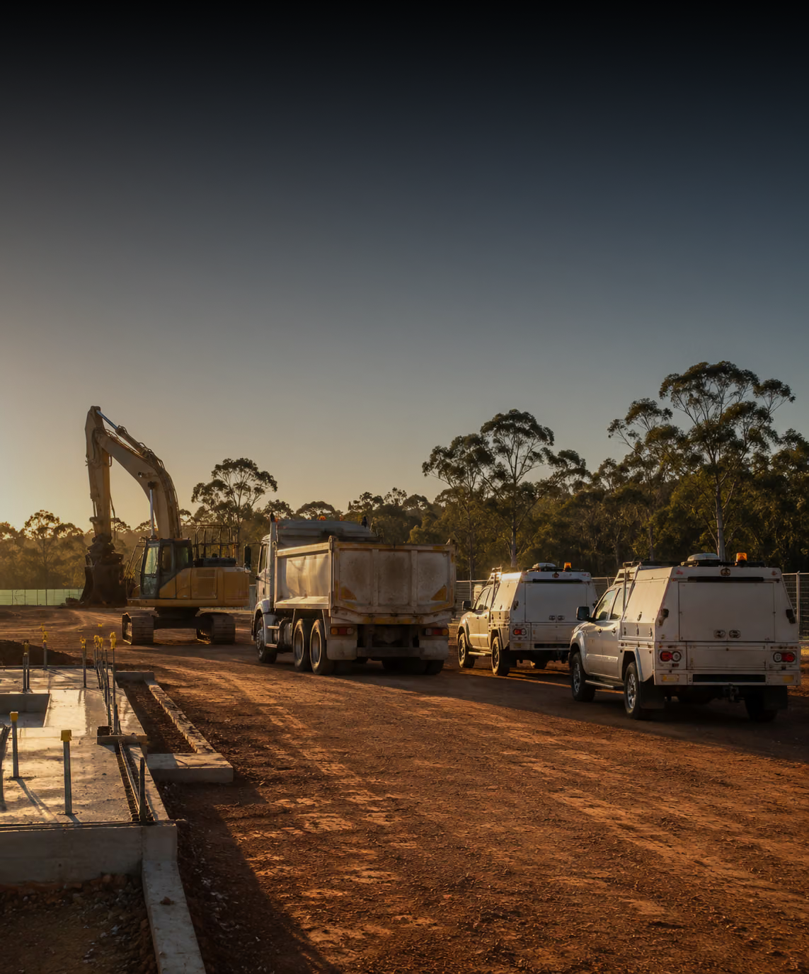 Photograph: a site fleet on a working construction site at first light. Dark and cool, room at the top, no text in the artwork.