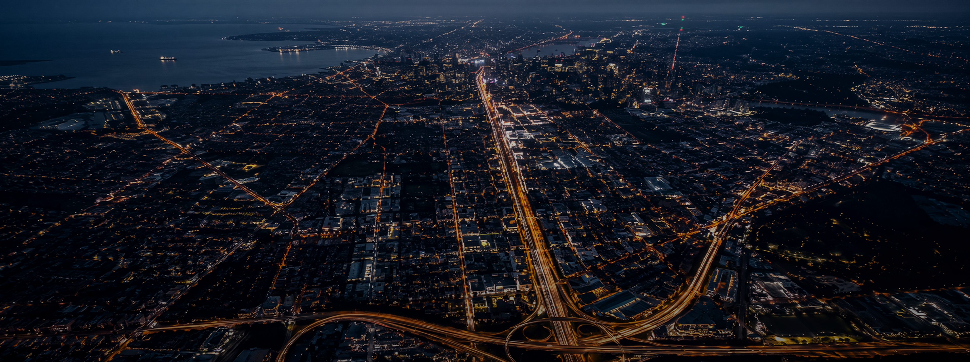 Full-bleed scene, edge to edge: a city at night from above with its arterials lit, dark and cool so the headline and the accordion read over it. No text in the artwork. 2400x1400.